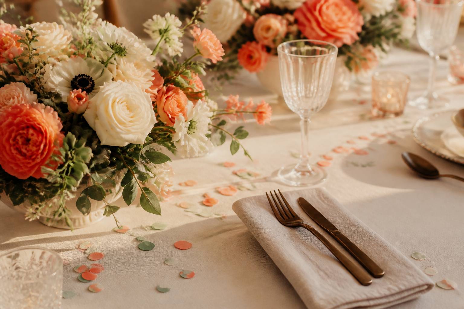 Détails d'une table de mariage avec des confetti, des fleurs corail et des couverts en laiton